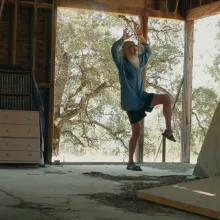 In an abandoned-looking shell of a house, a man with a long white beard does some stretches.