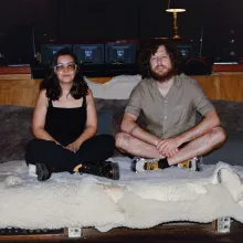 A brunette woman dressed in black sits next to a man with a beard on a couch covered in various furs. 