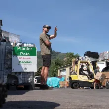 A man in a cap and sunglasses stands near a bin labeled “Planter Pots,” pointing into the distance. Behind him, a forklift carries crushed recyclables as two workers load boxes near a shipping container. The scene is set against a backdrop of trees, hills and a clear blue sky.