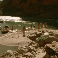 16mm still of a group of seven young adults and their canoes, on the banks of the Colorado River surrounded by canyons.
