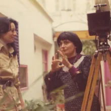 A candid shot of a woman in a khaki outfit with long brunette hair(Raganelli) standing next to a woman with a bowl cut(Agnes Varda) as they converse. 