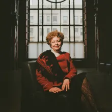 A woman wearing a vibrant red jacket sits regally in a chair.