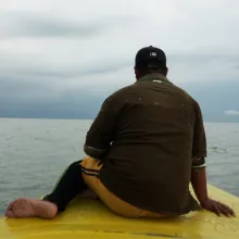 Barefoot and steady, a man sits on the bow of a yellow boat, his back to the camera, shirt weathered by time. He faces the horizon as the vessel cuts quietly through the water. The lake stretches endlessly under heavy clouds, evoking both solitude and endurance.