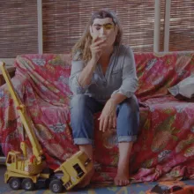 On a cloth-covered couch on an outdoor porch, a woman holds a Halloween mask over her face.