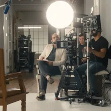 In a white studio, a white man wearing a white jacket sits in a chair, behind a camera and lights.