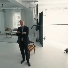 In a studio, Chris Hansen stands with his arms crossed, looking at a bearded white man in a white jacket.
