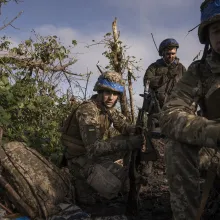 Three soldiers, with helmets and a small Ukrainian flag on their uniforms, crouching holding their guns outdoors