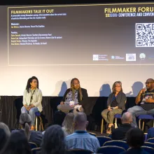 With a movie screen behind them that details the information for this Filmmaker Forum panel, four seated panelists with microphones face a crowd