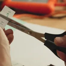 Closeup of a person cutting a piece of paper with "It was" written on it in half with a pair of black scissors.