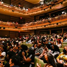 A crowded auditorium with green seats full of a clapping, sitting audience.