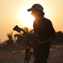 A man wearing a baseball cap and carrying a camera and tripod strolls through an arid landscape, illuminated by a sunset.