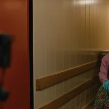 A camera on a tripod captures a portrait of a woman wearing a pink sweater, latex gloves, and a wooden indigenous mask in a nondescript room.