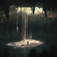 From far away, a film still of a man standing in a shaft of light in a vast underground cavern.