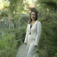 Woman with shoulder length brown hair in a white suit stands against a lush green garden backdrop.