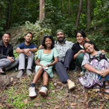 Six brown-skinned individuals (three men, three women) sit on a grassy knoll in a forest, smiling at the camera