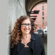 A vertical image of a smiling white woman with long curly brown hair, wearing glasses and a black shirt, leaning against a stone wall.