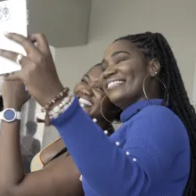 Vanessa and Tamara stand close together, smiling brightly as they take a selfie on a smartphone. Vanessa, in a royal blue top with long braids and hoop earrings, holds the phone out in front of them, while Tamara, dressed in a patterned outfit, leans in beside her. Both women radiate joy, their wide smiles filling the frame. Just behind them, Vanessa’s husband embraces someone tightly, his face pressed against a shoulder. The moment is tender yet bittersweet—Vanessa and Tamara are capturing one last photo t
