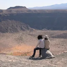 Two people sit on a rock against a vast desert landscape before them