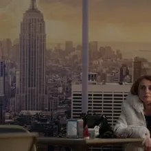A woman with shoulder-length auburn hair wearing a white winter coat sits alone at a table with a blown-up print of the New York City skyline behind her