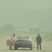 In a dusty road, two men with helmets on stand on either side of a broken down, battered car.