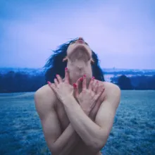 A still from the film shows Adelina standing alone in an open field, her head tilted upward toward the sky. Her bare shoulders and tightly crossed arms convey exposure and restraint, while her upward gaze suggests longing and a deep desire for freedom. The cold blue tones of the landscape and the distant city reinforce her emotional isolation.