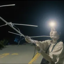 A young Asian woman stands in the middle of the street at night holding an antenna, lit only by the headlamp she sports on her head