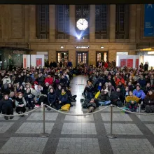 Hundreds of folks sit on the ground at Leipzig's Central Station, at a film screening; a projector can be seen behind them below a clock
