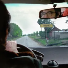 View from inside a car, a white man with dark hair and glasses can be seen driving through the rearview mirror as he passes a sign that reads Treblinka
