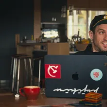 A white man wearing a cap and a hoodie sits at a kitchen table with his laptop, which is adorned by a communism sticker and another that reads "Let Cuba Live"