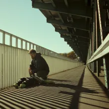 Wide shot of a bearded middle-aged white man with short-cropped hair kneeling over a mannequin of sorts at the railing on a bridge