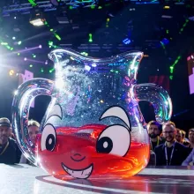 A CGI-rendered Kool-Aid-looking juice jug stands on a stage at a convention with men with lanyards looking in the audience