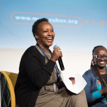 A Black woman with short-cropped hair sits at a stage next to another Black woman; both are holding mics and smiling widely