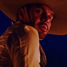 A brown-skinned young man wearing a mariachi suit and hat, lit by a red light looks down at the camera against a dark blue backdrop