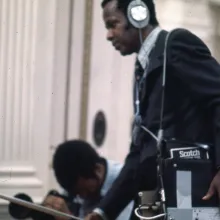 One man looks through a film camera viewfinder while another holds a microphone and sound recorder while wearing headphones.