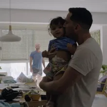 A young brown-skinned man hugs and kisses a young toddler girl amid a slightly messy living room while a balding white man in shirt and shorts looks at them in the background