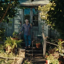 A man in blue jeans and a long sleaved shirt stands in front of the door of his house and his lush, green front yard.
