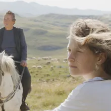A young white blond girl in the mountains is seen in profile, while an older white woman with her grey hair pulled back rides a white horse