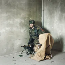 A soldier in uniform crouches inside an empty concrete room with a machine gun