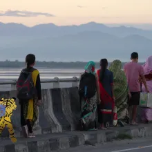 A group of men in colorful shirts and women in colorful saris walk along a sidewalk on a bridge, mountains in the distance, with an animated tiger with a backpack picking up the rear