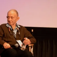 An older, balding white man with short wispy white hair wears a blue shirt and a brown cardigan while sitting on a director's chair on a stage, holding a mic and a water bottle.