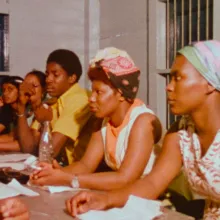 A group of Black women (in bright colorful sleeveless dresses and headwraps) sit at a table, as if in a meeting