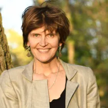 A woman with white skin smiles at the camera. She has short, curly salt-and-pepper hair, blue eyes, red lipstick, and is wearing a quilted black leather blazer. Headshot of Alison McAlpine