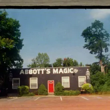 a 16mm film still of a low, black painted brick building with skeletons painted on the front labeled "Abbot's Magic" in large white lettering
