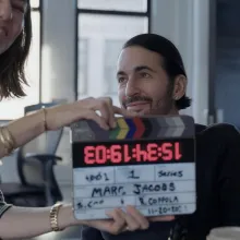 A middle-aged white man with slicked back black hair and five o'clock shadow sits in front of a camera as a white woman with shoulder-length brown hair holds a clacker that reads "Marc Jacobs" in front of him
