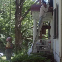 Two female sisters in their 60s and 70s look on as their brother fixes the siding of an old white house on a ladder.
