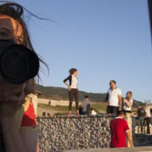 A white woman holding a camera is seen in a mirror as a group of people stand along in the background in a desert field