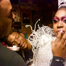 A man putting glitter on a drag queen while another man watches happily in background.