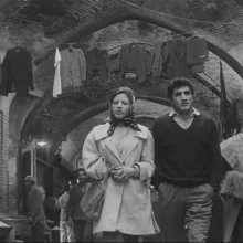 Black and white image of a woman in a coat and silk scarf over her hair walking alongside a man in pants ad a v-neck sweater around a Middle-Eastern market