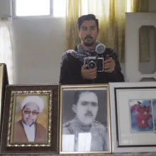 A young brown-skinned man with short black hair and goatee beard holds a camera in front of a mirror where we can see various photos of older Middle-Eastern men in frames.