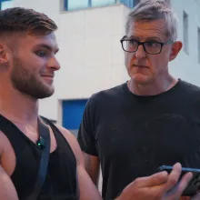 A middle-aged white man in a black t-shirt and rimmed glasses stares incredulously at a young muscled white man in a black tank top as he stares into his smartphone